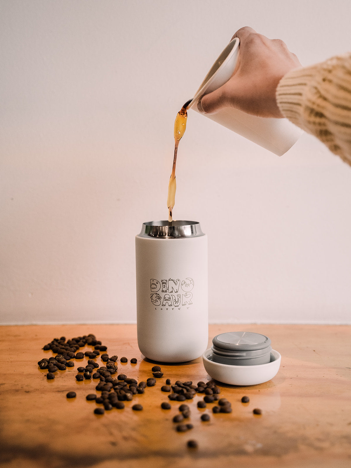 An out of frame hand pouring coffee into a tall white travel mug with the logo for "dinosaur coffee" printed on the mug. Coffee beans are scattered to the front left side of the tumbler.