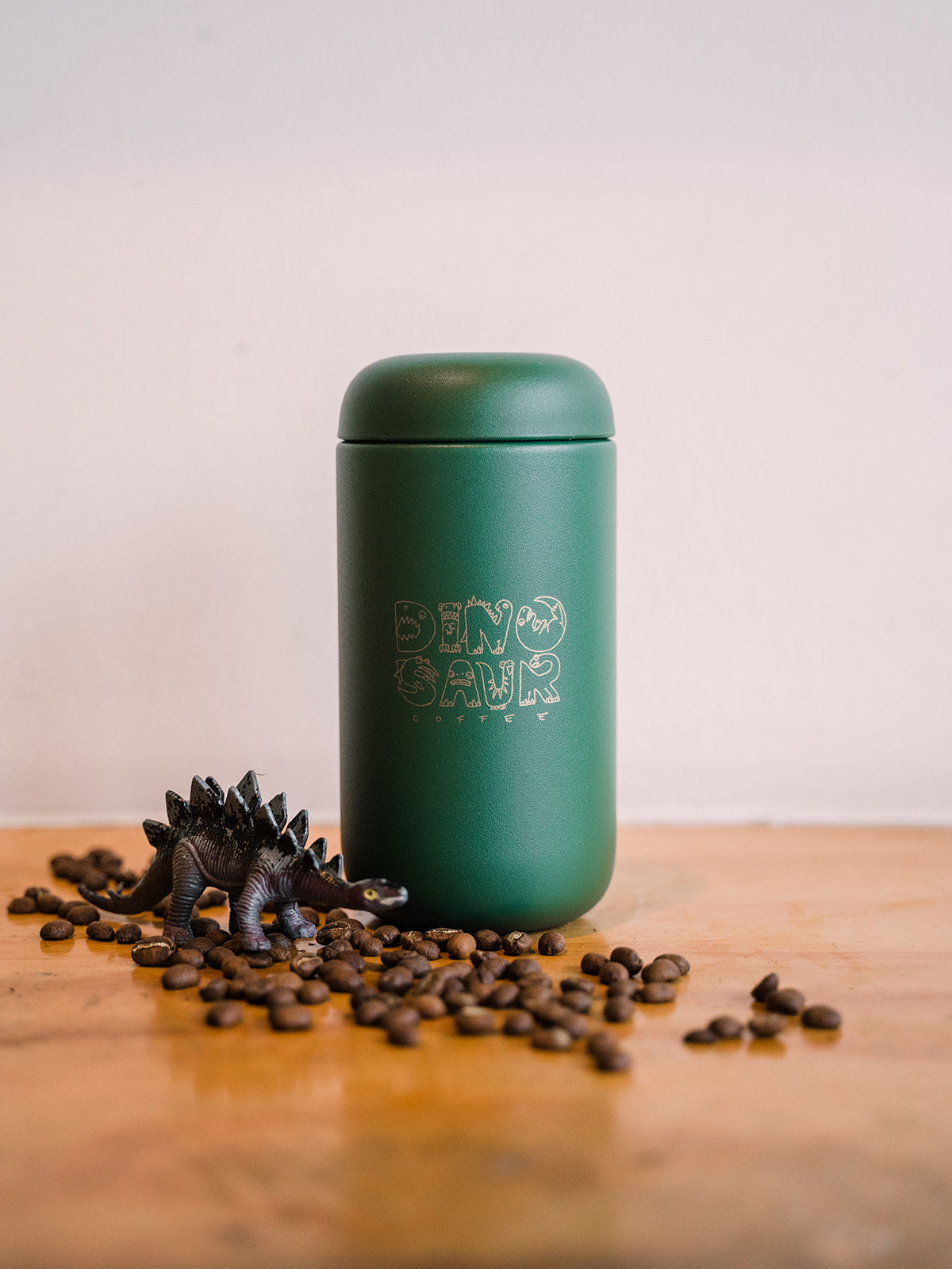 A green travel tumbler that reads "dinosaur coffee" surrounded by a toy dinosaur figure sitting over some coffee beans