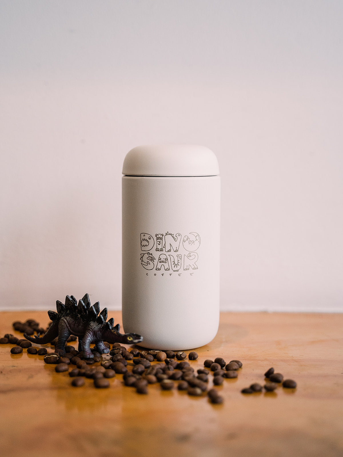 A white travel tumbler that reads "dinosaur coffee" surrounded by a toy dinosaur figure sitting over some coffee beans