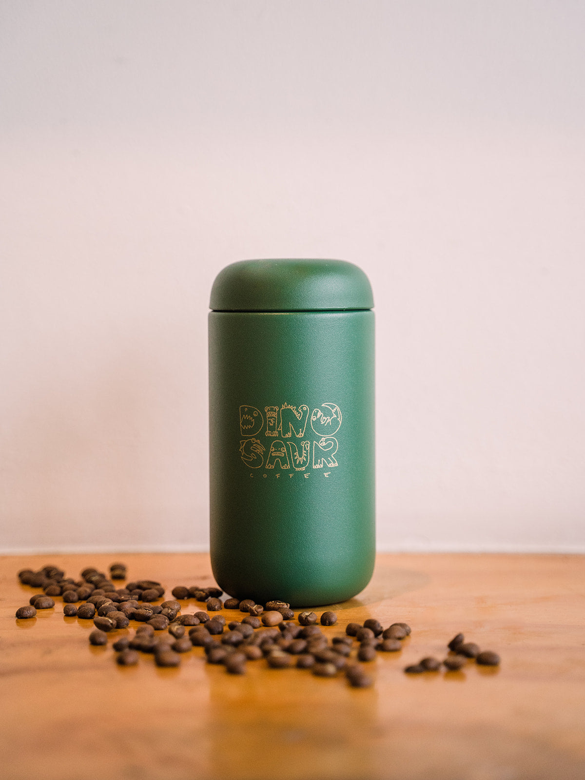 A green travel tumbler that reads "dinosaur coffee" surrounded by coffee beans