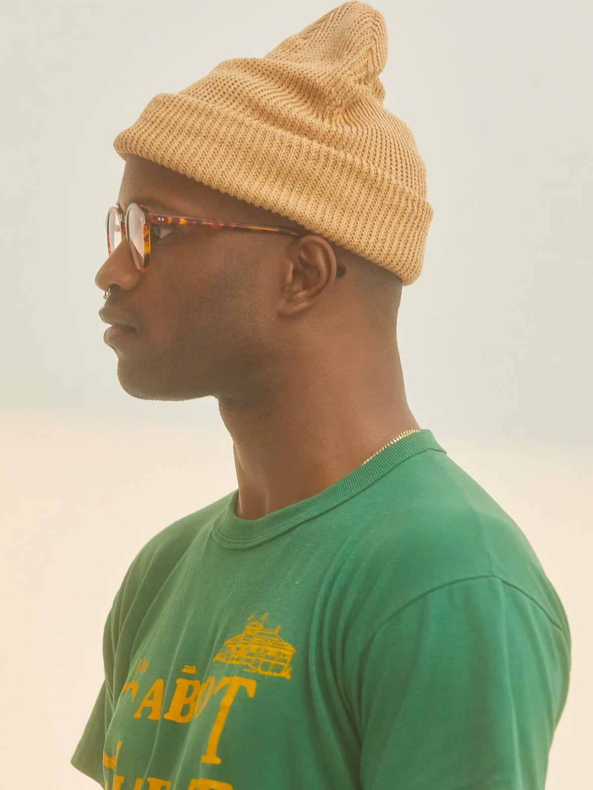 A profile shot of a man in a green shirt wearing a tan-colored knit beanie