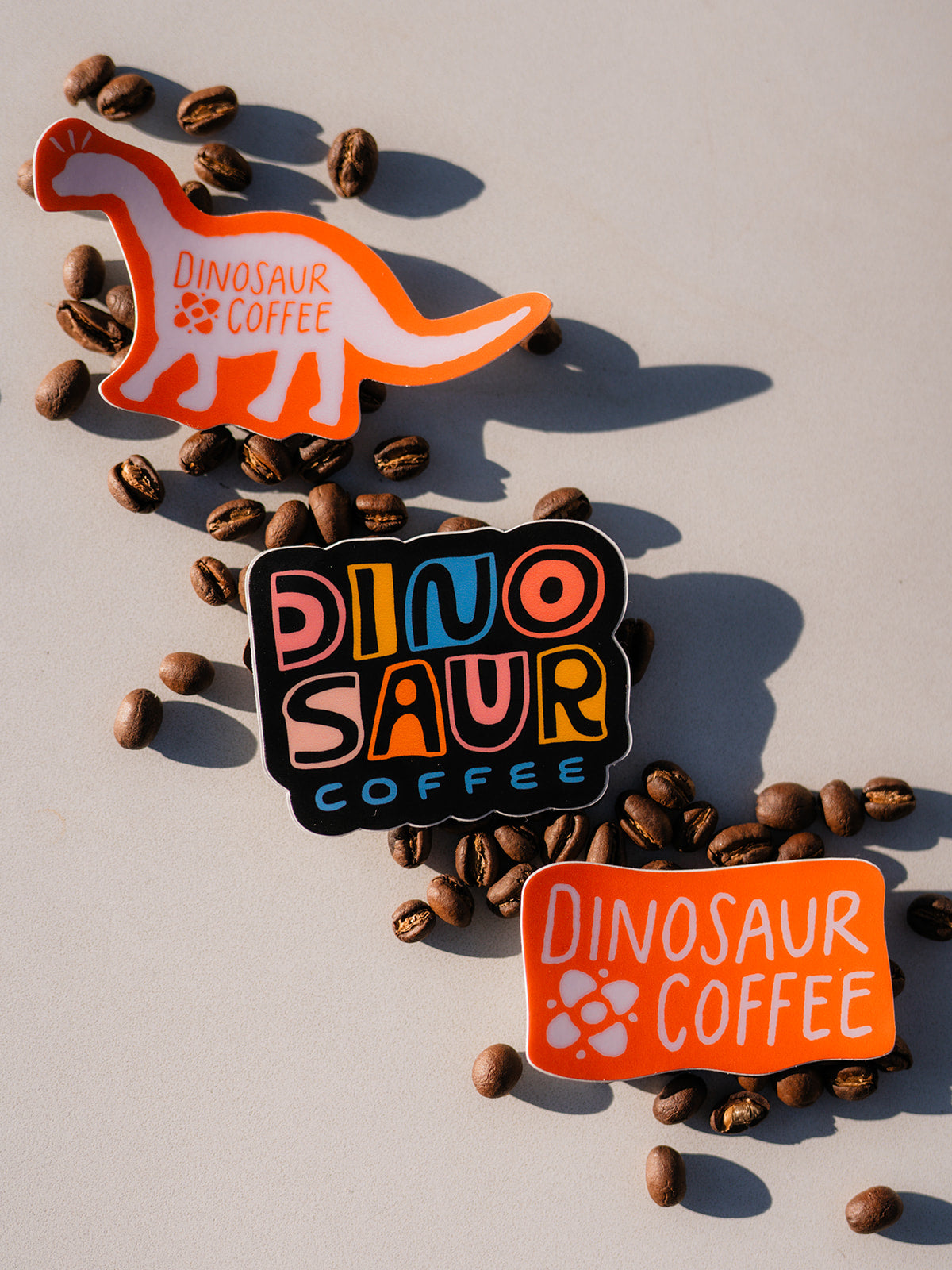 A close-up of 3 colorful stickers in a diagonal arrangement over a scattering of coffee beans. The top left corner sticker is of a pink silhouette with an orange border of a dinosaur with a logo that reads "dinosaur coffee". The middle sticker is carved-style colorful lettering on black that reads "DINOSAUR COFFEE". The bottom right sticker is an orange rectangle with pink lettering that reads "DINOSAUR COFFEE"