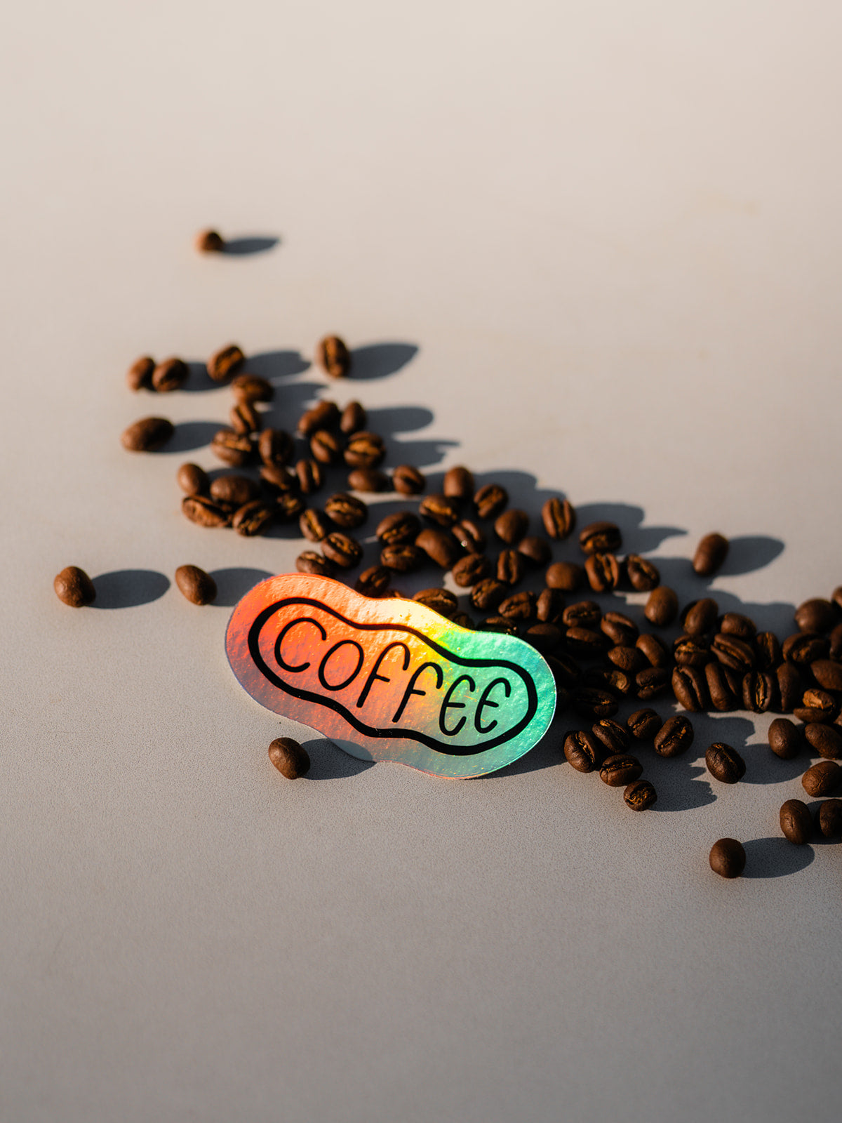 A close-up view of a rainbow sticker that reads "coffee" . It is set on top of a scattering of coffee beans. The background is white