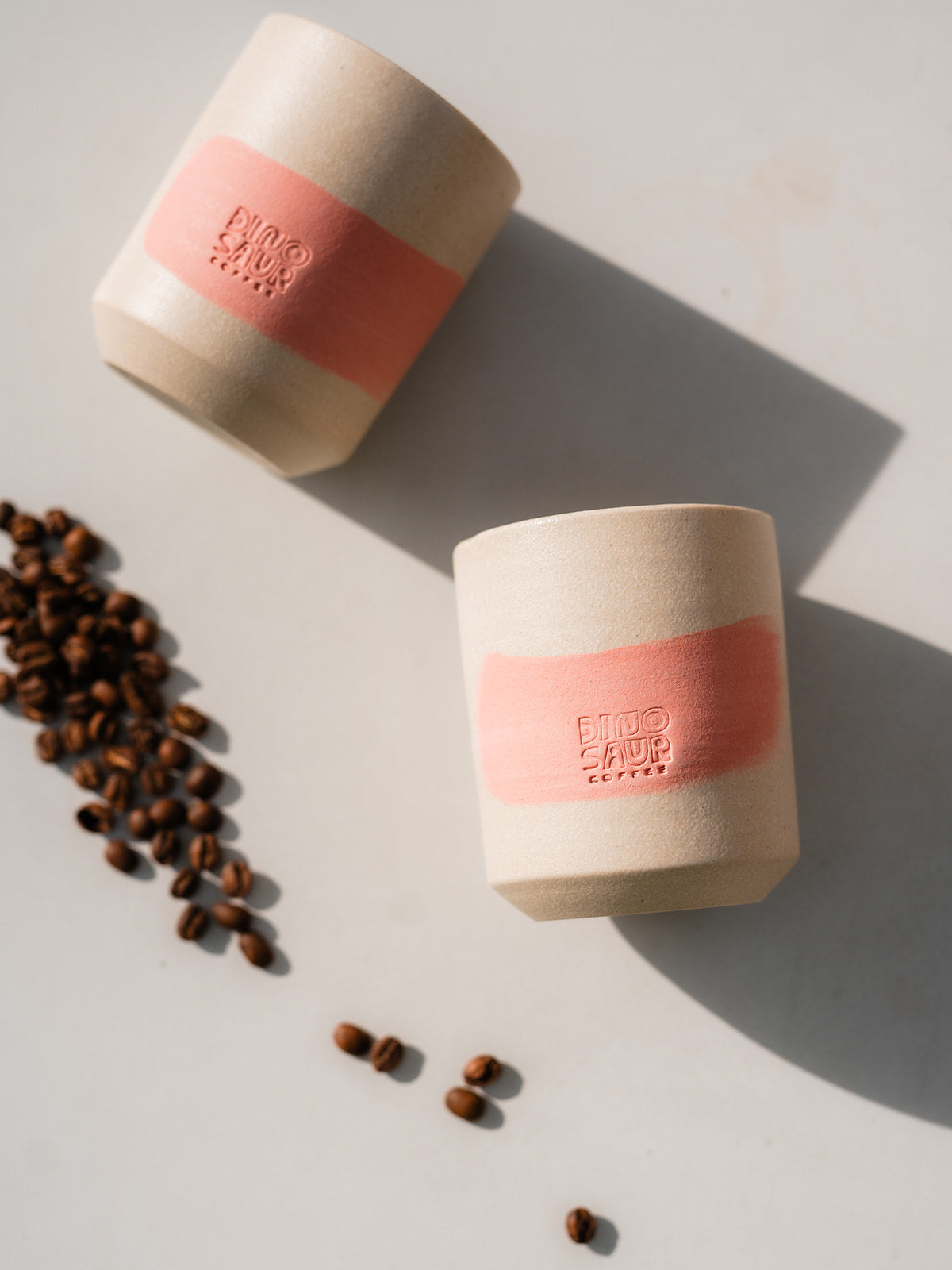 Two of the same cream-colored cups, both with a pink brushstroke lined on the left side with a small pile of coffee beans