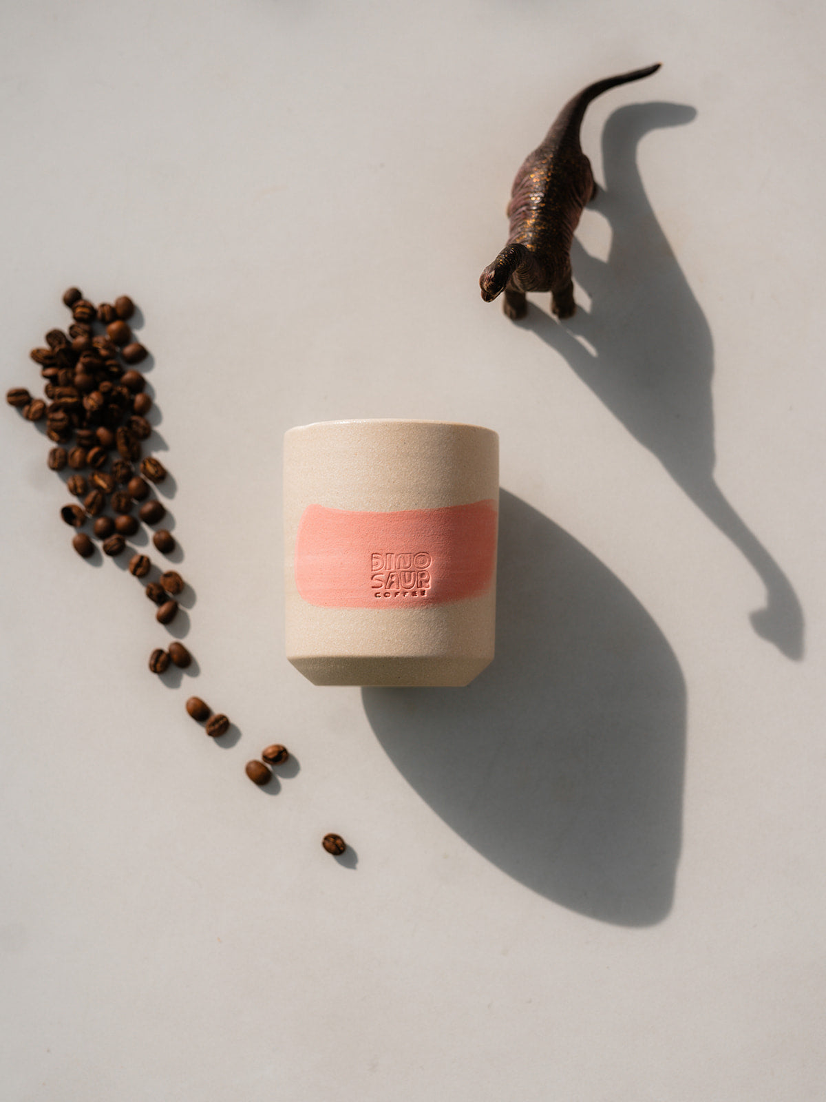 A cream colored cup with a pink brushstroke surrounded by a toy dinosaur and a small pile of coffee beans