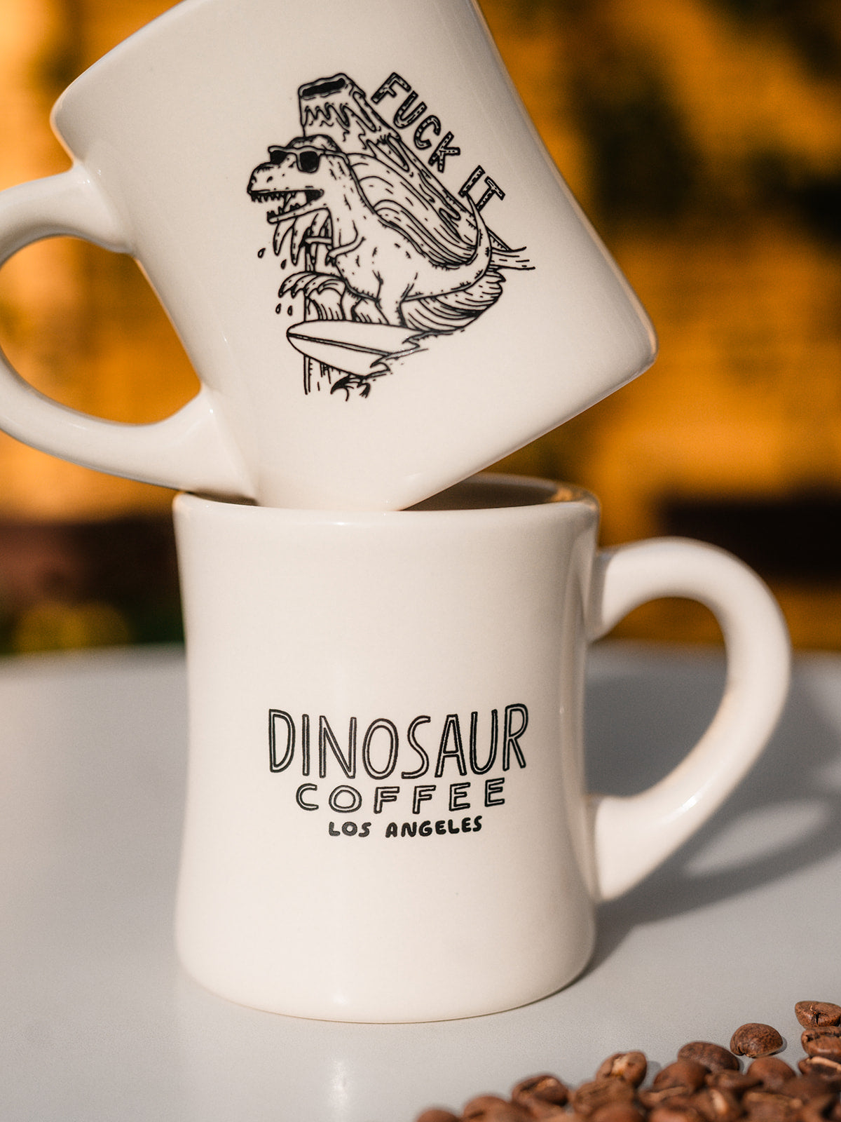 Two white mugs stacked one over the other. The mugs are decorated with black line-art. The top mug is an illustration of a surfing dinosaur with the words "FUCK IT" drawn beside and erupting volcano. The bottom mug says "dinosaur coffee los angeles"