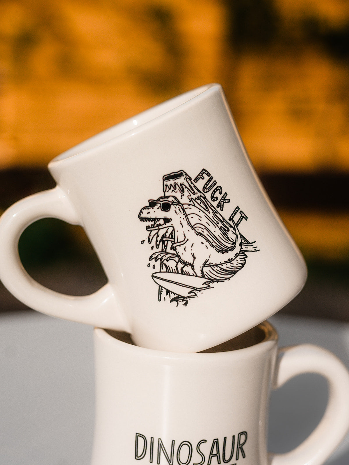 A white diner mug resting inside of another. The mugs are decorated with black line-art. The top mug is an illustration of a surfing dinosaur with the words "FUCK IT" drawn beside and erupting volcano. The bottom mug reads "DINOSAUR"