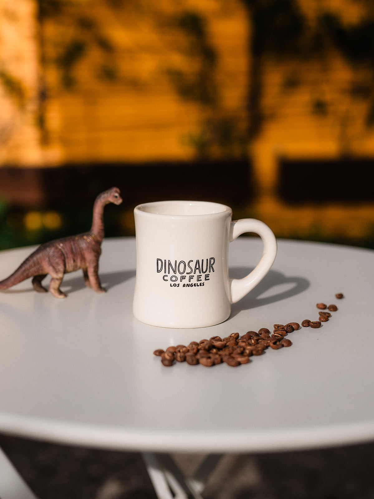 A photo of a white diner mug sitting in the middle of a white table surrounded by coffee beans and a toy dinosaur figure. The coffee mug reads "dinosaur coffee los angeles"