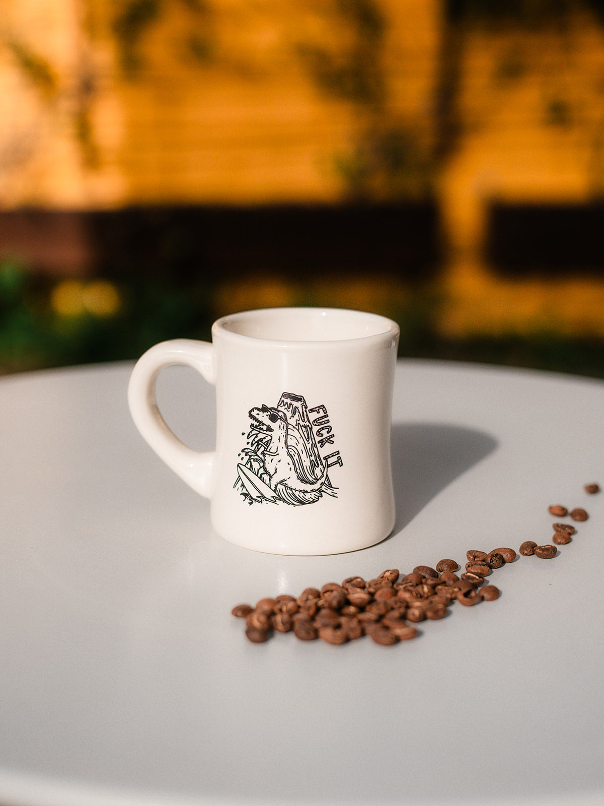 A photo of a white diner mug sitting in the middle of a white table surrounded by coffee beans. The coffee mug has a black line illustration of a volcano-surfing, sunglass-wearing dinosaur and the words "fuck it"