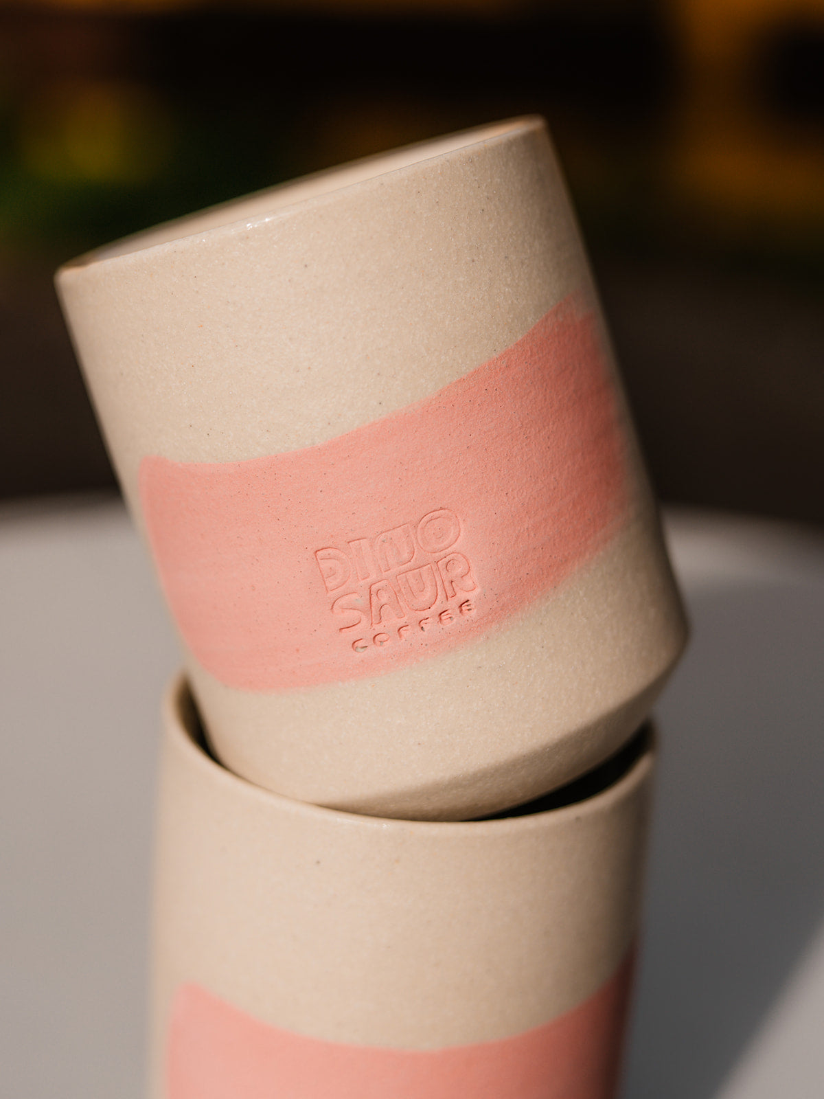 A close-up of a cream colored cup with a pink brushstroke and a stamp of Dinosaur Coffee engraved over the pink.
