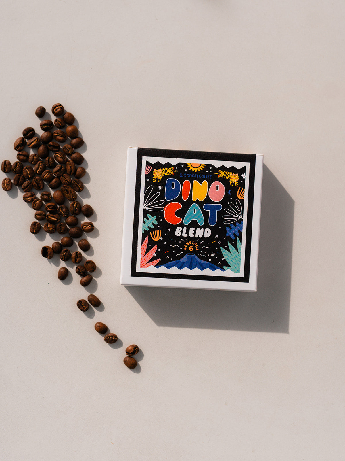 A white box laid over a white background. The box says "DINOCAT BLEND" in colorful bubble lettering. A small pile of coffee beans are set off to the left side of the box.