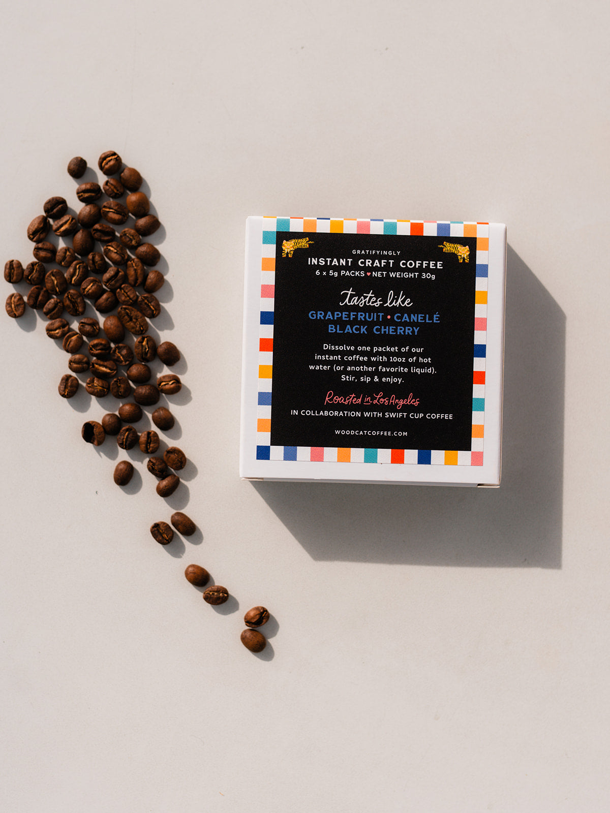 A white box laid over a white background. The box says "Instant craft coffee. 6x 5g packs. Net weight 30g. Tastes like: grapefruit, canéle, black cherry. Dissolve one packed of our instant coffee with 10oz of hot water (or another favorite liquid). Stir, sip & enjoy. Roasted in Los Angeles. In Collaboration with Swift Cup Coffee. Woodcatcoffee.com". A small pile of coffee beans are set off to the left side of the box.