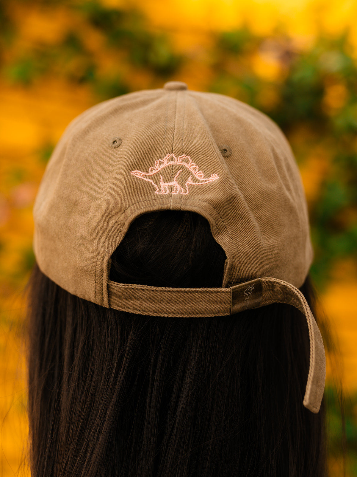 A close up of the back side of an olive green baseball cap with a pink embroidery of a dinosaur