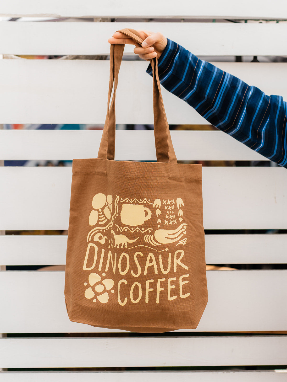 A close-up of brown tote bag printed with yellow icons and "DINOSAUR COFFEE". A person is holding the strap of the tote with their index and middle fingers.