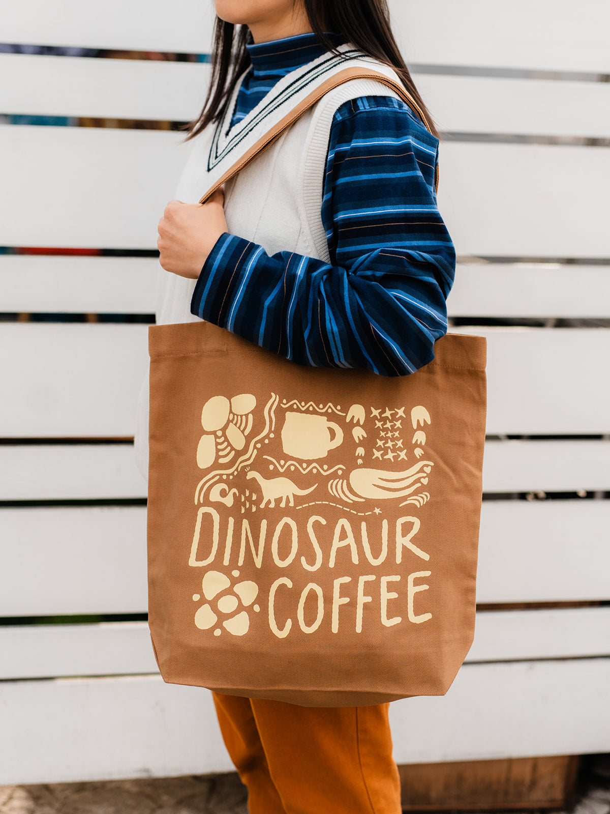A close-up of brown tote bag printed with yellow icons and "DINOSAUR COFFEE". The tote is hanging from a person's left shoulder as they hold on to the strap with their hand.