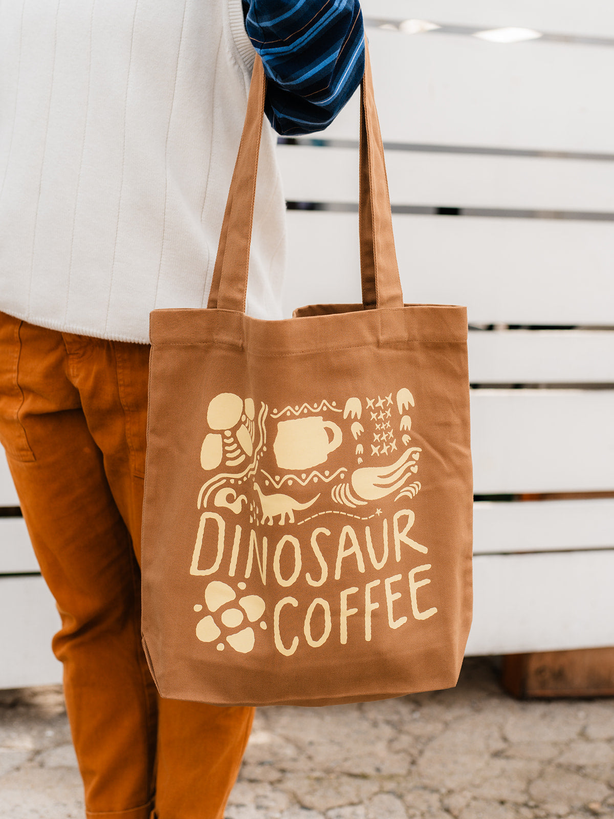 A close-up of brown tote bag printed with yellow icons and "DINOSAUR COFFEE". The tote is dangling from the crook of a person's arm