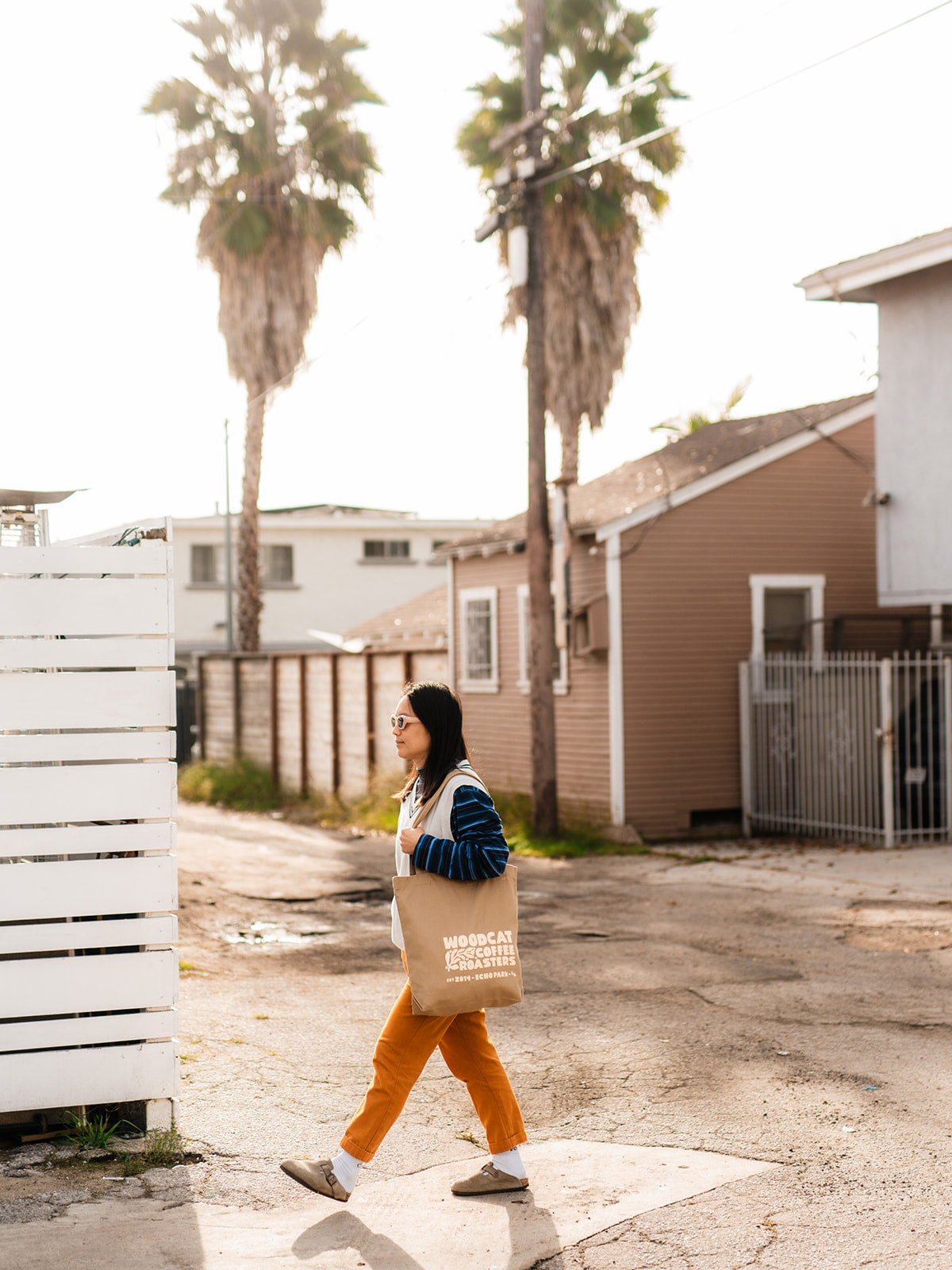 A brown haired woman walking through an alley with a brown tote bag that reads "woodcat coffee roasters" slung over her left shoulder