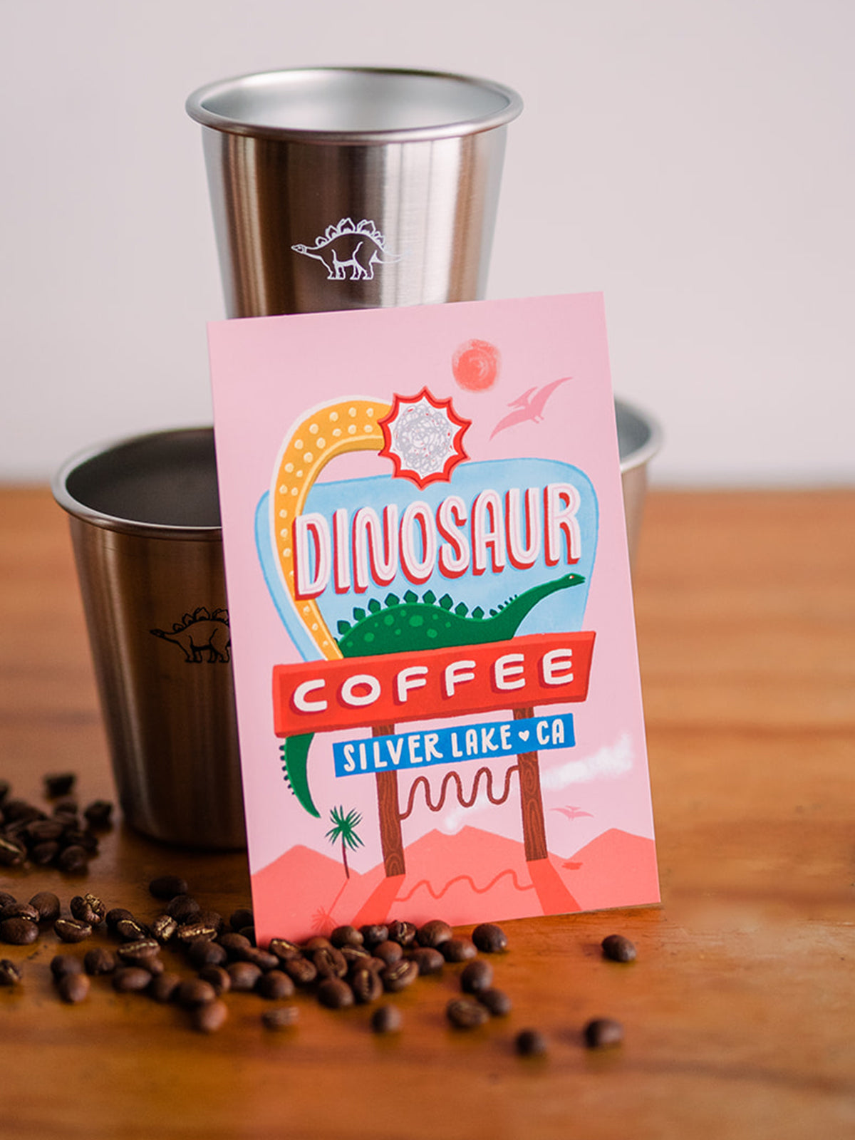 A close-up of a rectangular postcard with a vintage sign illustration of a desert-like scene that reads "Dinosaur Coffee Silver Lake CA". It is leaning against 2 tin cups with small brontosaurus logos printed on them. There are coffee beans scattered to the left side of the postcard just off to the side in the foreground.