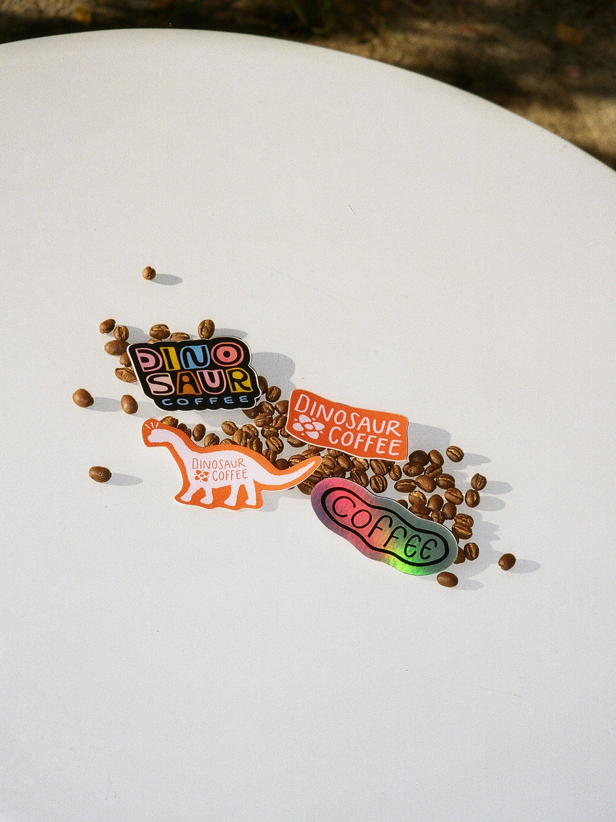 A distant photo of a round white table with an assortment of 4 colorful stickers, 3 of which read "DINOSAUR COFFEE" and one that reads "COFFEE" in black lettering on an iridescent rainbow background. The 4 stickers lie on top of a scattering of coffee beans.