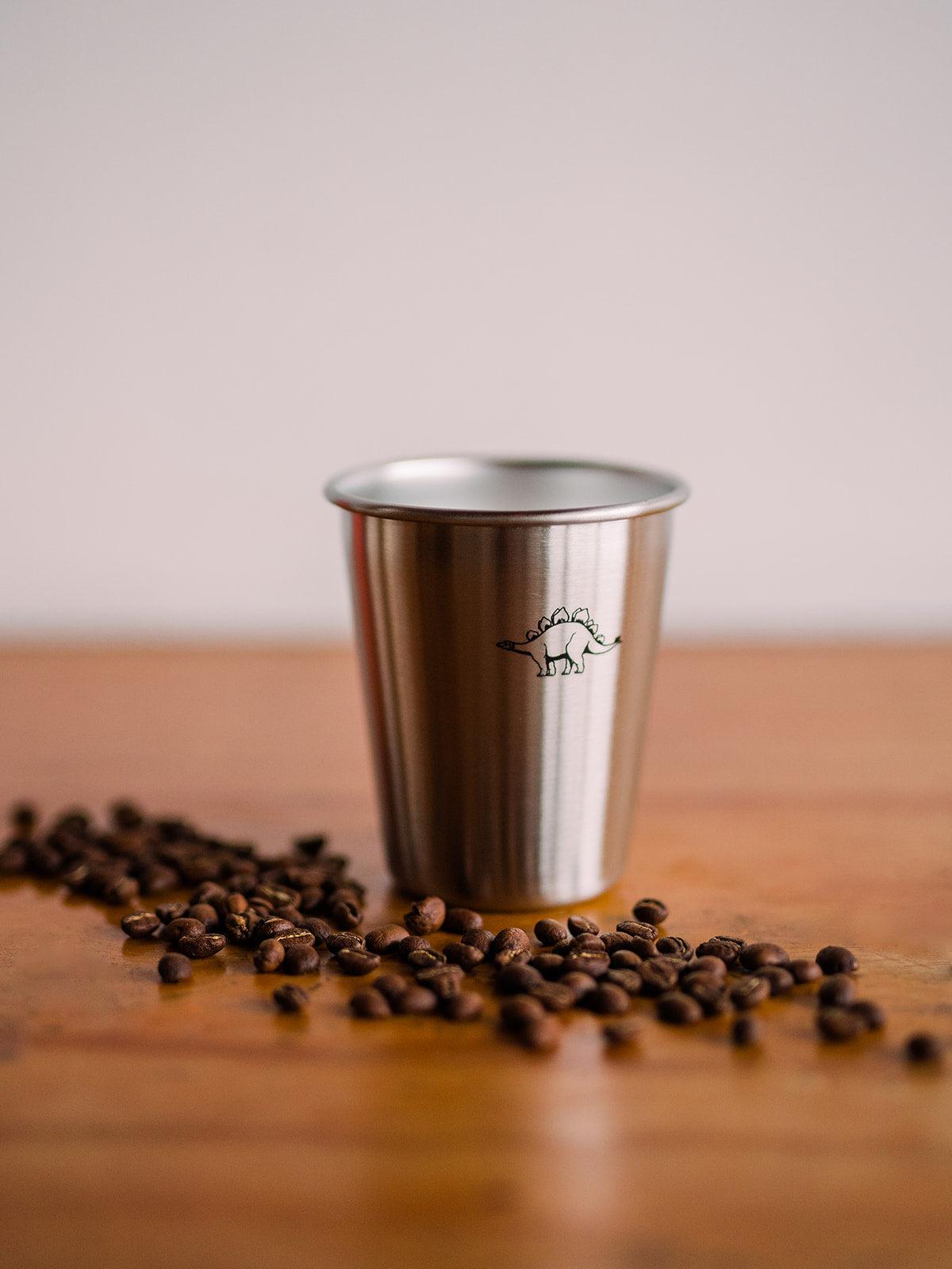 A close up of a tin cup with a black dinosaur printed on it. Coffee beans are scattered in front of the cup over a wooden surface.
