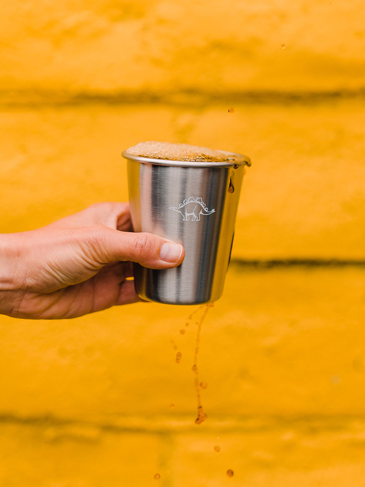 A close up of a hand in front of a yellow wall holding a tin cup filled with coffee dripping out of one side.