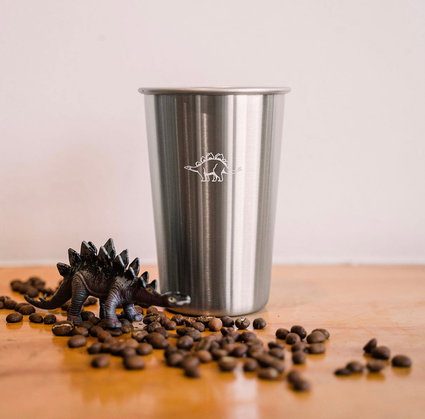 A close up of a tall tin cup printed with a white dinosaur. Toward the left side sits a toy dinosaur figurine surrounded by a scattering of coffee beans