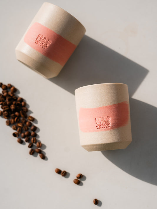 Two of the same cream-colored cups, both with a pink brushstroke lined on the left side with a small pile of coffee beans