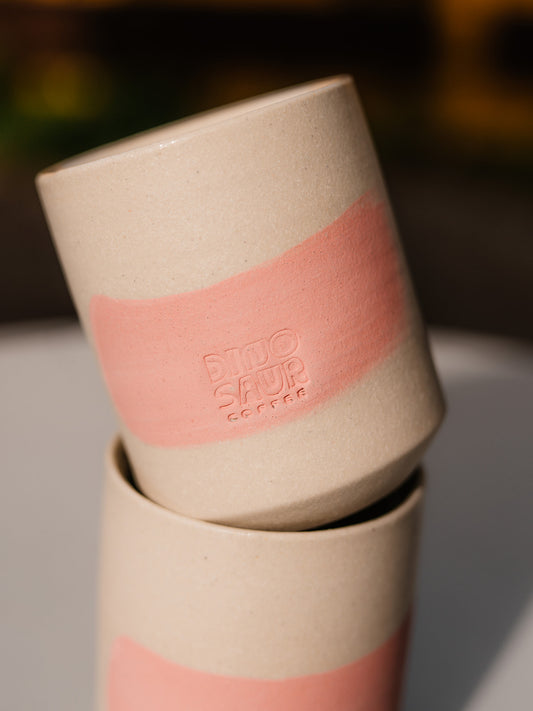 A close-up of a cream colored cup with a pink brushstroke and a stamp of Dinosaur Coffee engraved over the pink.