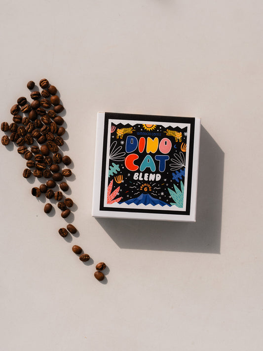 A white box laid over a white background. The box says "DINOCAT BLEND" in colorful bubble lettering. A small pile of coffee beans are set off to the left side of the box.