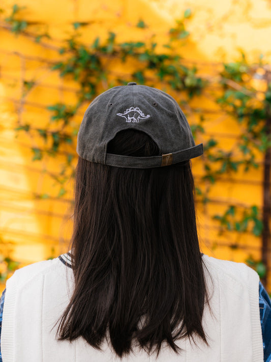 A photo of the back side of an gray baseball cap with a blue embroidery of a dinosaur