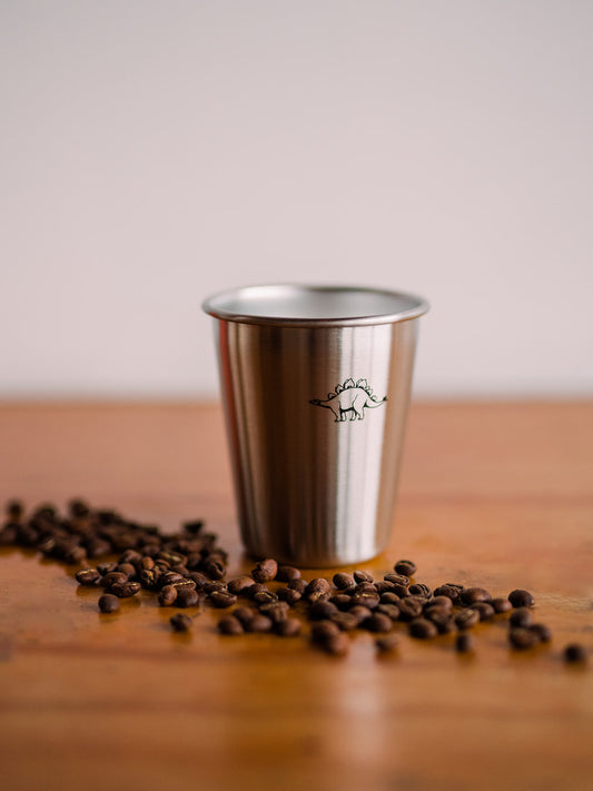 A close up of a tin cup with a black dinosaur printed on it. Coffee beans are scattered in front of the cup over a wooden surface.